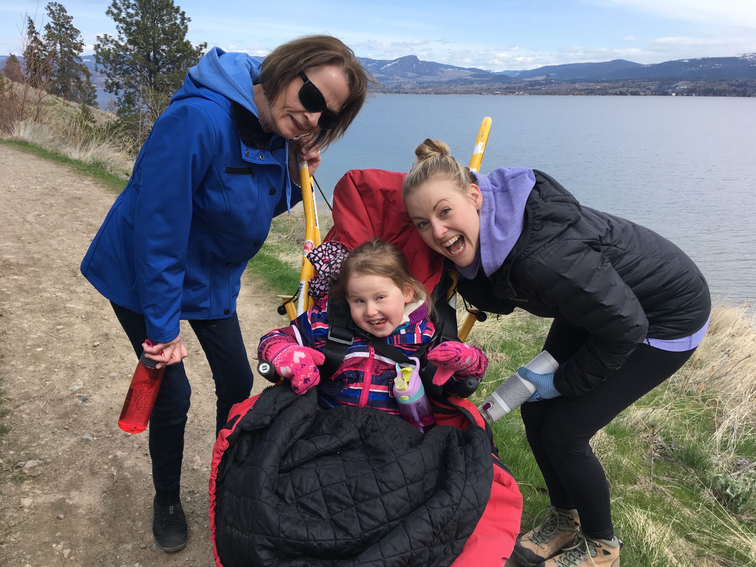 A young girl in a yellow youth trailrider. She is in a red sleeping bag and making a silly face. beside her and leaning over her are two women, one with blonde hair and the other with brunette. They are on a dirt path, behind them is a lake and mountains.