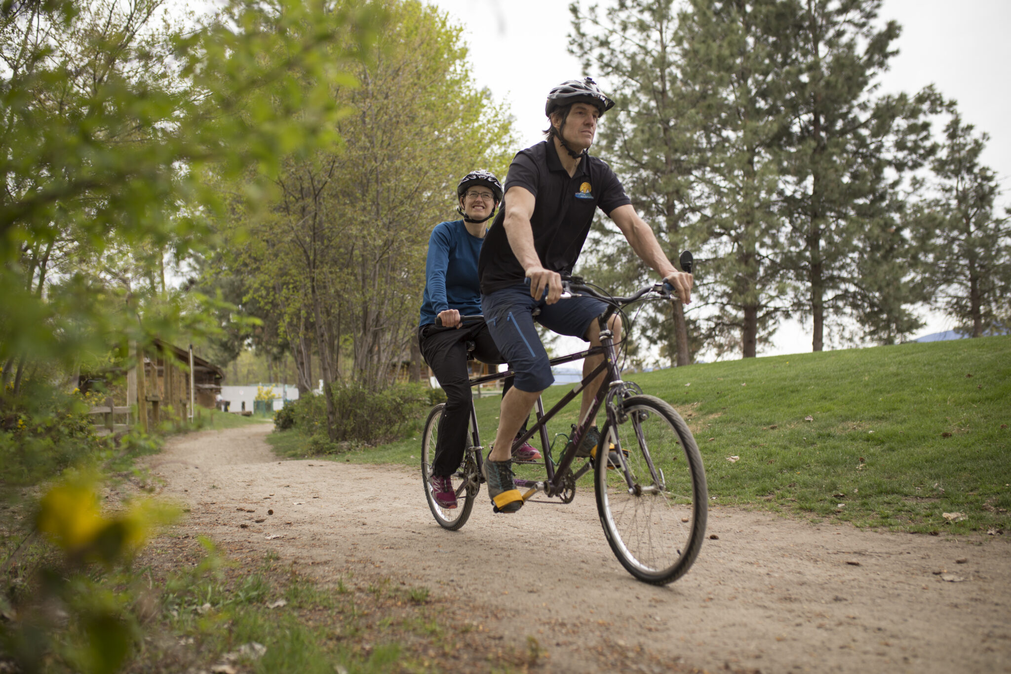a man and a woman using a dark purple tandem upright cycle on a forest path.