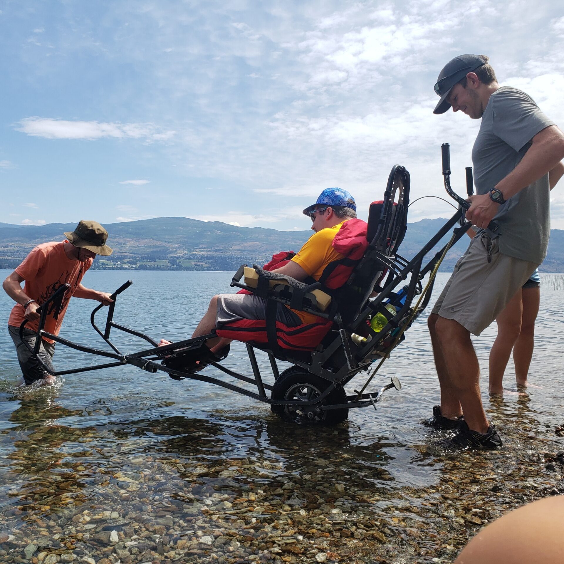 a man using a trail rider - and adaptive hiking chair on the shoreline being balanced by two volunteers.