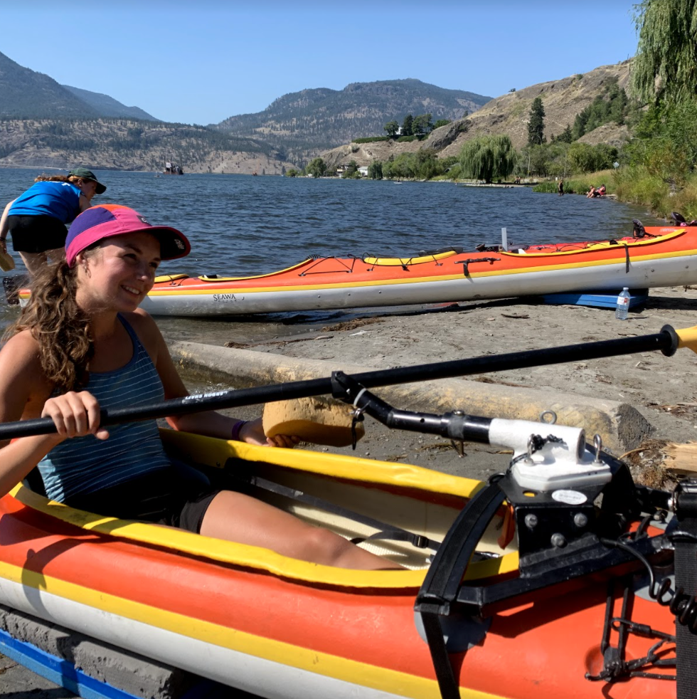 A woman in a kayak on the shore, demonstrating the one-armed rigger.