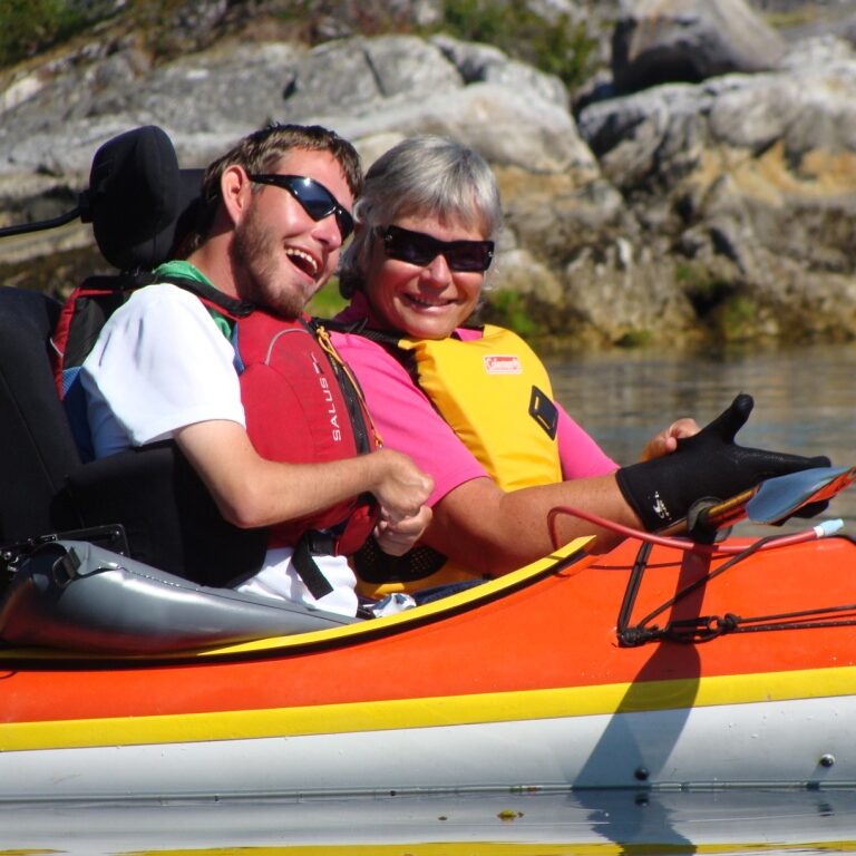 A man and a woman in separate kayaks. The man is being handed a black paddle. The man is seated in black supportive seating, similar to that of a wheelchair.