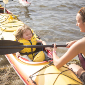 A woman handing a young girl in a kayak a paddle. The kayak is in the water.