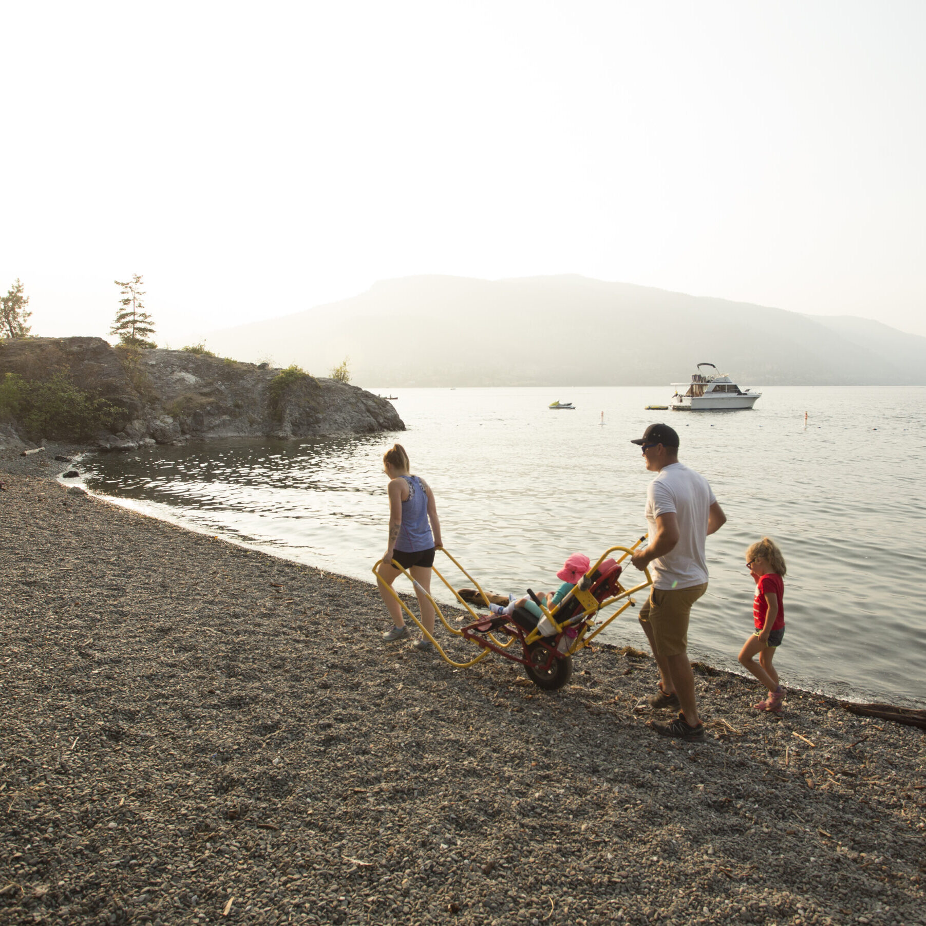 A beach scene. A woman is pulling a yellow youth trailrider while a man pilots it. There is a child seated in the trailrider while another child follows behind.