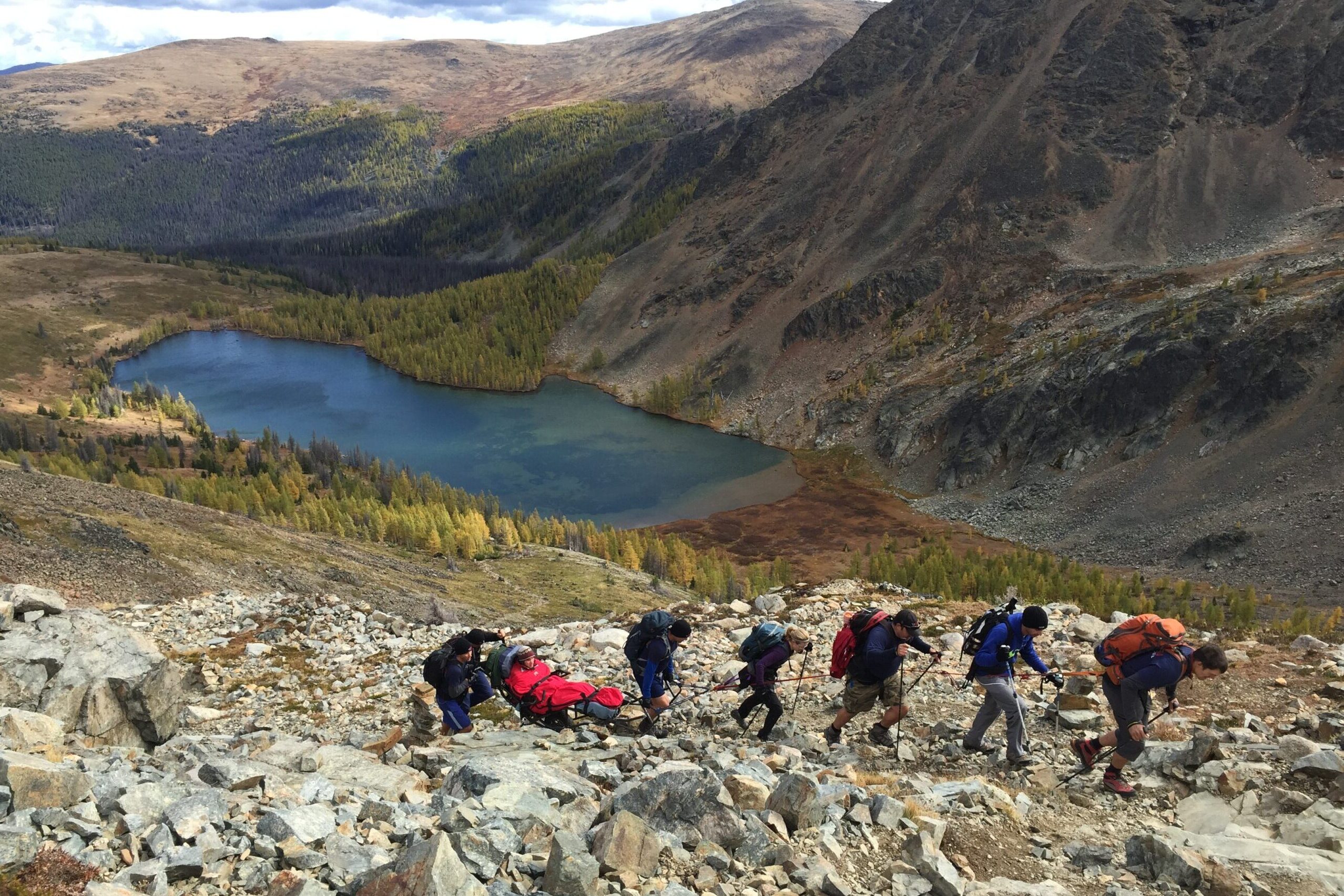 Seven hikers in rocky terrain. One is using a trailrider. There is a blue lake in the background.